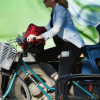 Eine seitliche Aufnahme eines Fahrrads, das eine Frau in Richtung des linken Bildrandes fährt. Davor steht ein schwarzer Stuhl, im Hintergrund ist ein grüner sportstättenartiger Platz zu sehen.