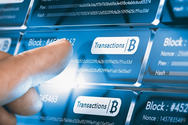 Glaskacheln mit weißer Schrift vor blauem Hintergrund. Die Wörter "Block" und "Transactions" sind zu erkennen, daneben ist ein Bitcoin-Zeichen. Davor ist eine Hand zu sehen, ein Finger deutet auf die Kachel in der Mitte.