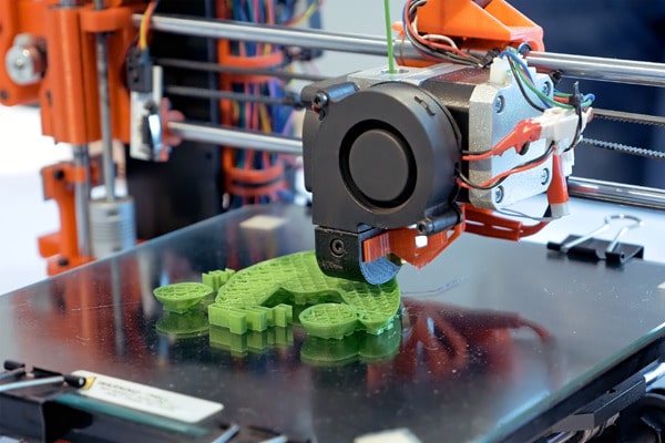 Zu sehen ist ein 3D-Drucker bei der Arbeit. Unter dem Druckkopf liegt ein grünes Gebilde auf dem Tisch