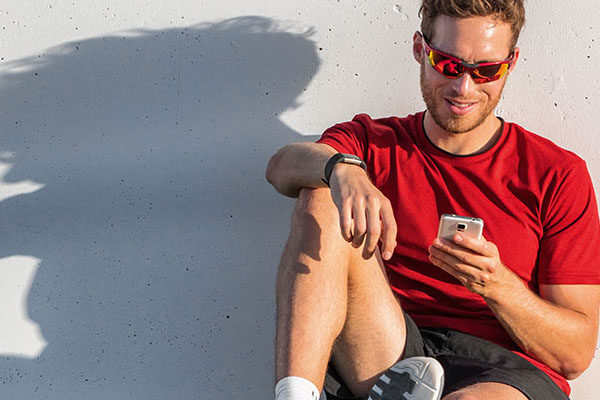 Ein Mann in Fitness-Kleidung sitzt mit einem Smartphone in der Hand vor einer Mauer. Auf die Mauer fällt sein Schatten.